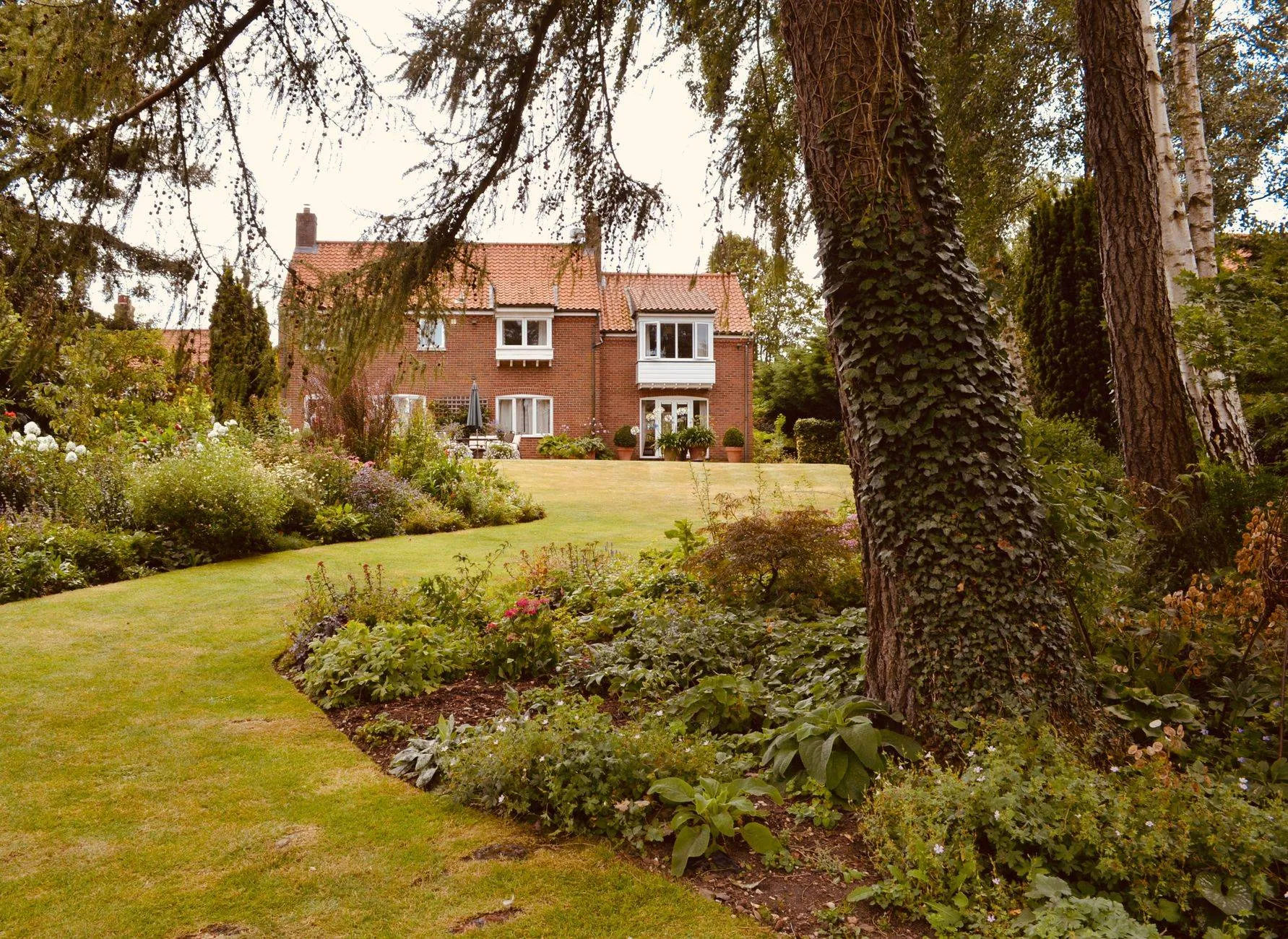 A curving lawn edged by a deep border under mature trees, a brick house beyond