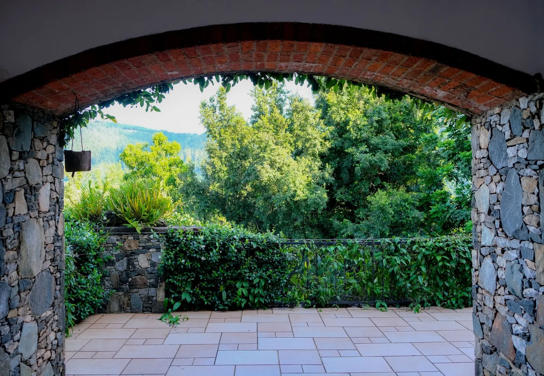 A pale sandstone terrace seen through a stone archway, hedges and trees beyond